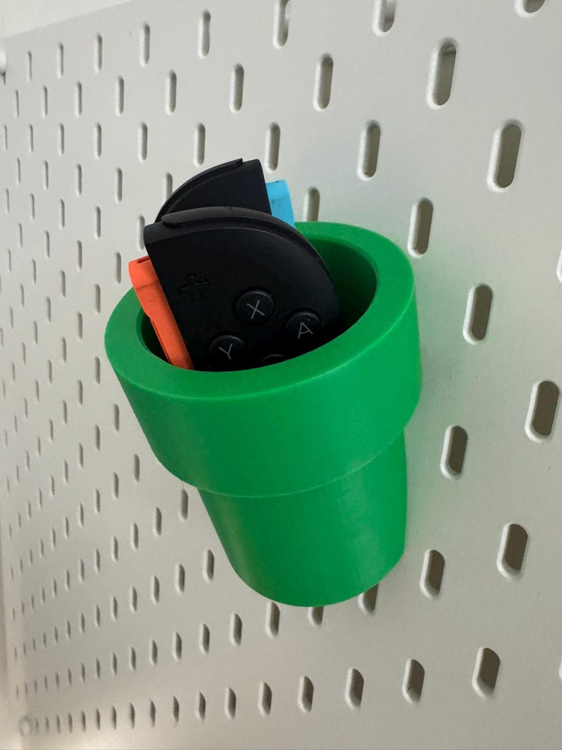 May include: A bright green, 3D-printed storage container shaped like a pipe, holding black, orange, and blue game controllers. The container is mounted on a white pegboard with evenly spaced holes.