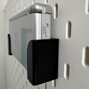 Op de afbeelding: Een zilveren handheld-spelconsole met een zwarte wandbevestiging. De console heeft een aan/uit-knop met het label "POWER".