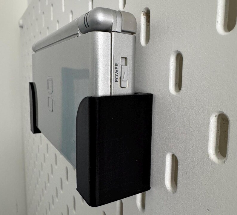 Op de afbeelding: Een zilveren handheld-spelconsole met een zwarte wandbevestiging. De console heeft een aan/uit-knop met het label "POWER".