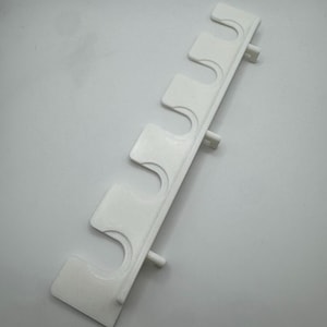 May include: A white, 3D-printed plastic wall mount with five curved slots for holding items. The mount has a rectangular shape with small cylindrical pegs extending from the back for attachment. The design is simple and functional.