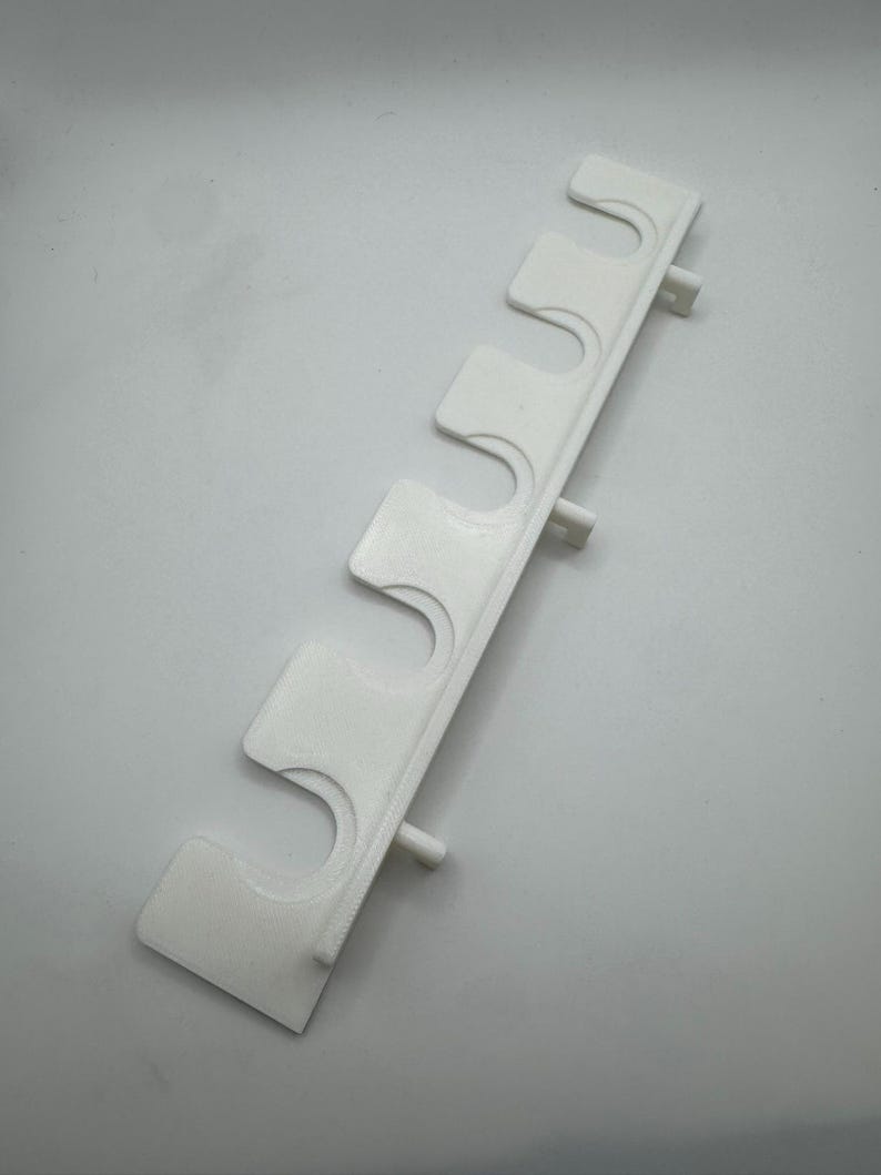 May include: A white, 3D-printed plastic wall mount with five curved slots for holding items. The mount has a rectangular shape with small cylindrical pegs extending from the back for attachment. The design is simple and functional.