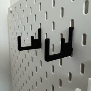 以下が含まれることがあります： 白いペグボードに取り付けられた黒いペグボードフック。フックはU字型で、アイテムを保持するように設計されています。ペグボードには、フック配置用の穴が均等に配置されており、多用途な収納ソリューションを提供します。