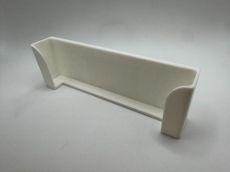 Pu&ograve; includere: Un contenitore bianco rettangolare con la parte superiore aperta, un bordo curvo su un lato e lati angolati. Realizzato in materiale opaco, poggia su una superficie bianca. Il design &egrave; semplice e moderno.