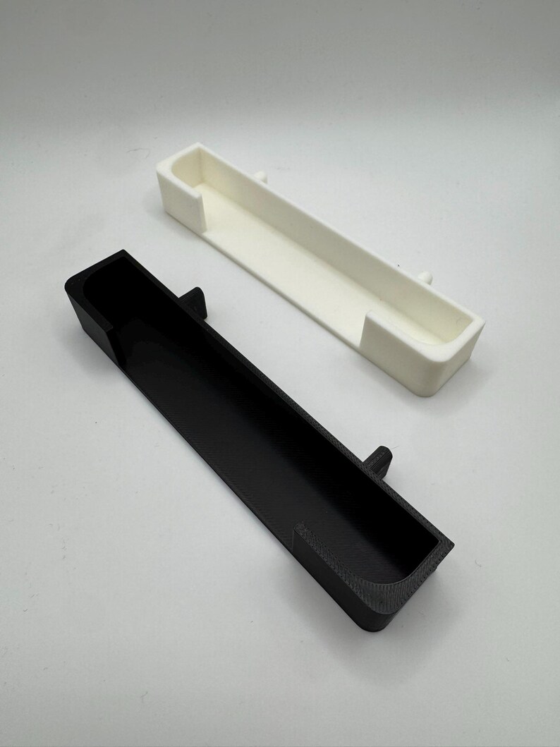 May include: Two rectangular, open-top containers. One is black, the other is white. Both have a rectangular shape with a small, raised tab on the side. The containers are likely 3D-printed.