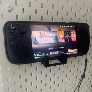 K&ouml;nnte beinhalten: Ein schwarzes Handheld-Gaming-Ger&auml;t mit einem Bildschirm, auf dem das Spiel "Hogwarts Legacy" angezeigt wird. Das Ger&auml;t ist mit einer schwarzen Halterung an einer Wand befestigt. Auf dem Bildschirm sind auch die Spiele "Plate Up", "Skyrim" und "No Man's Sky" zu sehen.