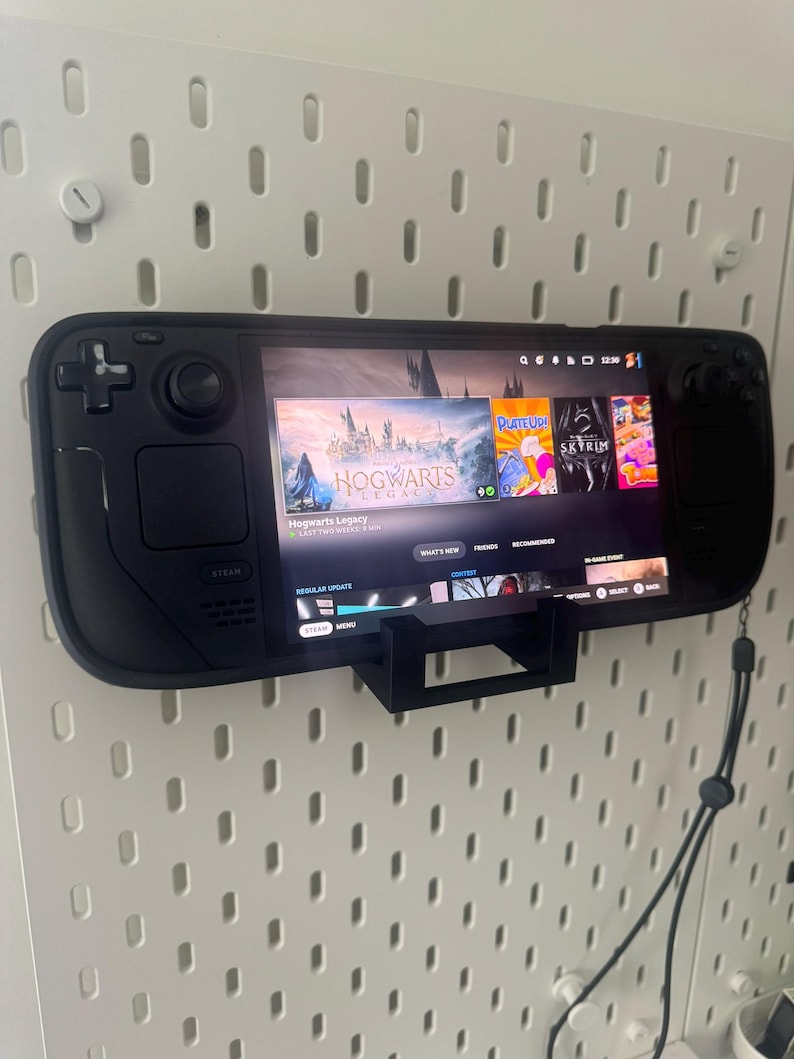 K&ouml;nnte beinhalten: Ein schwarzes Handheld-Gaming-Ger&auml;t mit einem Bildschirm, auf dem das Spiel "Hogwarts Legacy" angezeigt wird. Das Ger&auml;t ist mit einer schwarzen Halterung an einer Wand befestigt. Auf dem Bildschirm sind auch die Spiele "Plate Up", "Skyrim" und "No Man's Sky" zu sehen.