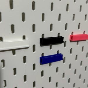 Pode incluir: Quatro organizadores de parede de pl&aacute;stico de diferentes cores, branco, preto, azul e rosa, s&atilde;o montados em uma placa de pinos branca.