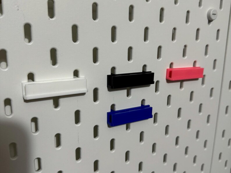 Pode incluir: Quatro organizadores de parede de pl&aacute;stico de diferentes cores, branco, preto, azul e rosa, s&atilde;o montados em uma placa de pinos branca.
