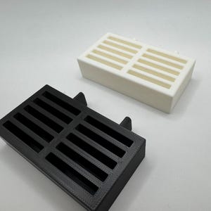 Op de afbeelding: Twee rechthoekige, sleufvormige ventilatieafdekkingen. E&eacute;n is zwart, de andere is wit. Beide hebben een reeks parallelle sleuven over de bovenkant. De zwarte afdekking heeft twee kleine lipjes aan &eacute;&eacute;n kant.
