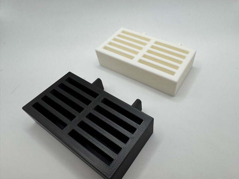 Op de afbeelding: Twee rechthoekige, sleufvormige ventilatieafdekkingen. E&eacute;n is zwart, de andere is wit. Beide hebben een reeks parallelle sleuven over de bovenkant. De zwarte afdekking heeft twee kleine lipjes aan &eacute;&eacute;n kant.
