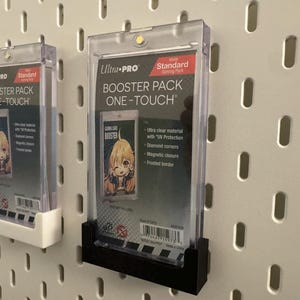 以下が含まれることがあります： 2つの透明なUltra-Pro One-Touch Booster Packケース。それぞれに漫画の画像が入ったトレーディングカードが入っています。ケースは白いペグボードに取り付けられています。1つのケースは白いホルダーに、もう1つは黒いホルダーに入っています。