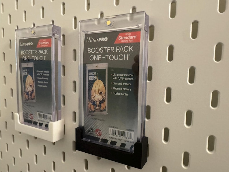以下が含まれることがあります： 2つの透明なUltra-Pro One-Touch Booster Packケース。それぞれに漫画の画像が入ったトレーディングカードが入っています。ケースは白いペグボードに取り付けられています。1つのケースは白いホルダーに、もう1つは黒いホルダーに入っています。