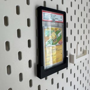 May include: A black display frame holding a Pokemon trading card, mounted on a white pegboard wall. The card features a Pikachu illustration and text. A white pegboard accessory is to the right of the frame.
