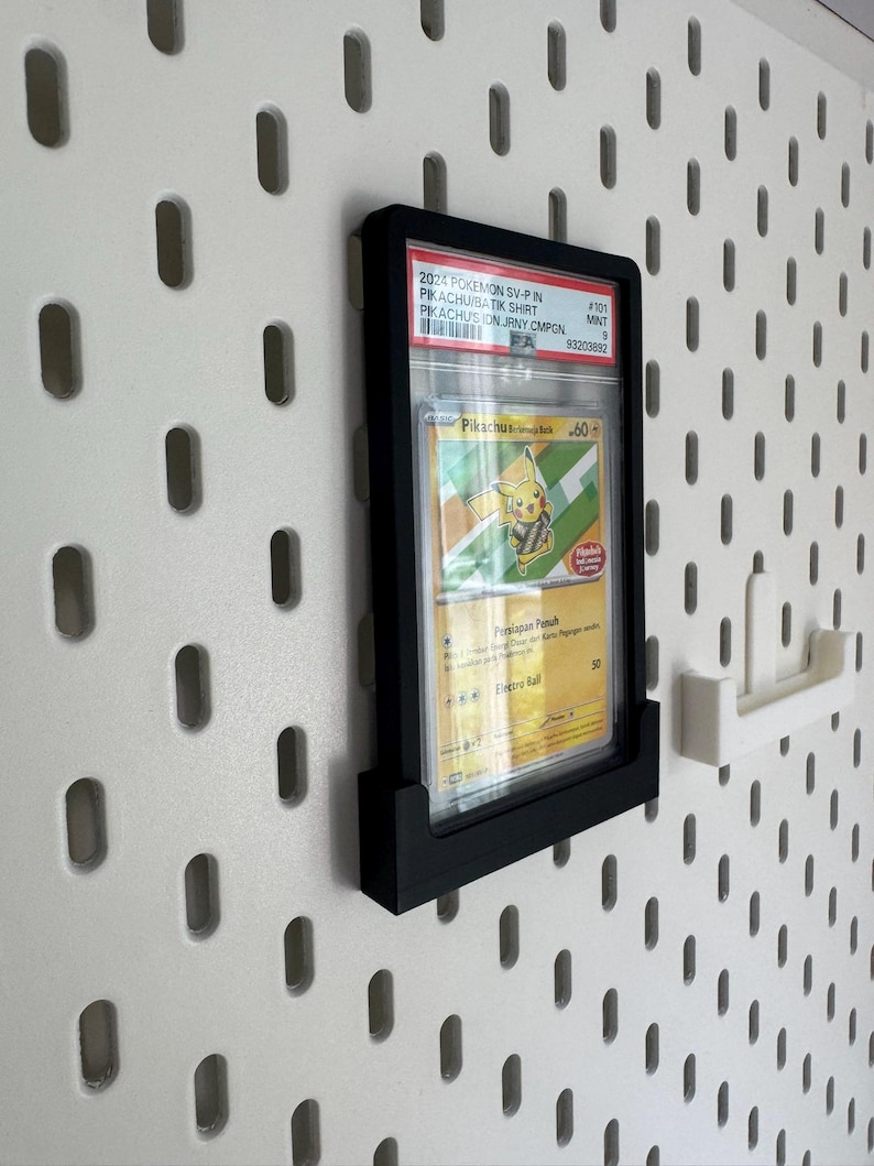May include: A black display frame holding a Pokemon trading card, mounted on a white pegboard wall. The card features a Pikachu illustration and text. A white pegboard accessory is to the right of the frame.