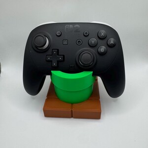 K&ouml;nnte beinhalten: Ein schwarzer Nintendo Switch Pro Controller auf einem braunen und gr&uuml;nen 3D-gedruckten St&auml;nder, der einem Super Mario Bros. Rohr und einer Ziegelplattform &auml;hnelt. Der Controller hat wei&szlig;e Akzente und verschiedene Tasten.