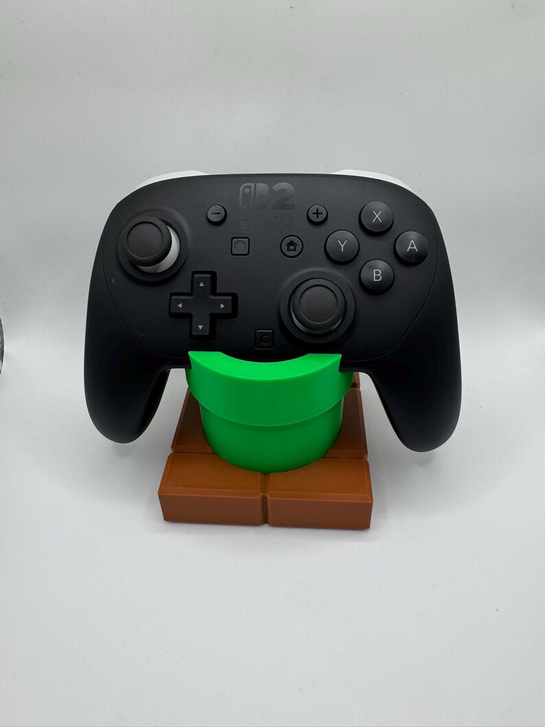 K&ouml;nnte beinhalten: Ein schwarzer Nintendo Switch Pro Controller auf einem braunen und gr&uuml;nen 3D-gedruckten St&auml;nder, der einem Super Mario Bros. Rohr und einer Ziegelplattform &auml;hnelt. Der Controller hat wei&szlig;e Akzente und verschiedene Tasten.