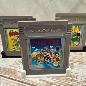 May include: Three gray Nintendo Game Boy cartridges. The center cartridge displays "Super Mario Land" artwork. The other two cartridges feature "Mario" and "Golf" game labels. All cartridges are in protective holders, showcasing classic video game titles.