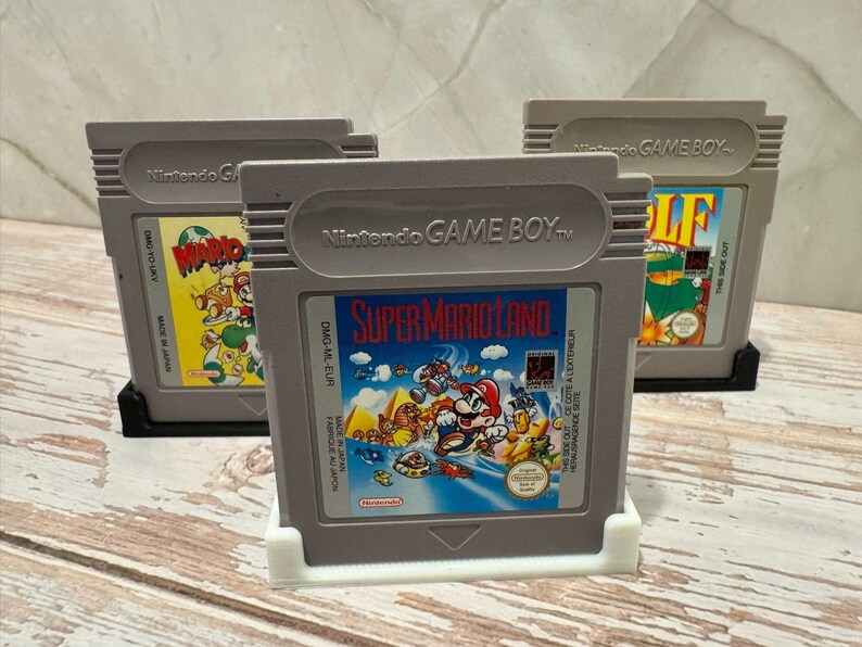 May include: Three gray Nintendo Game Boy cartridges. The center cartridge displays "Super Mario Land" artwork. The other two cartridges feature "Mario" and "Golf" game labels. All cartridges are in protective holders, showcasing classic video game titles.