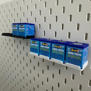 K&ouml;nnte beinhalten: Mehrere blaue Revell Aqua Color Farbt&ouml;pfe, die jeweils 18 ml Acrylfarbe enthalten, sind auf einer wei&szlig;en Lochwand angeordnet. Die Beh&auml;lter sind auf schwarzen und wei&szlig;en Regalen platziert. Der Text "Aqua Color" ist sichtbar.