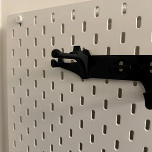May include: A white pegboard with a black accessory rail and hooks. The pegboard has evenly spaced, oval-shaped holes. The black rail has a series of hooks designed to hold items. A white screw secures the pegboard to the wall.