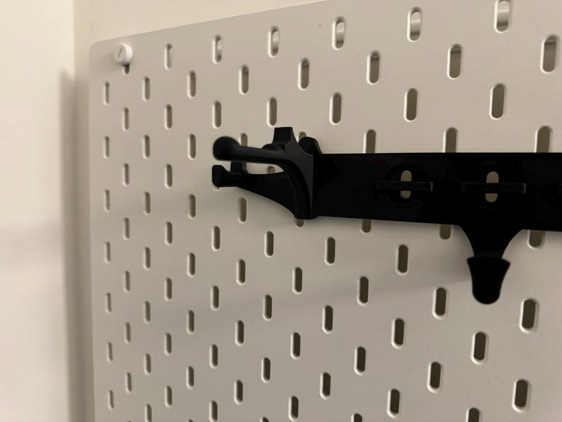 May include: A white pegboard with a black accessory rail and hooks. The pegboard has evenly spaced, oval-shaped holes. The black rail has a series of hooks designed to hold items. A white screw secures the pegboard to the wall.
