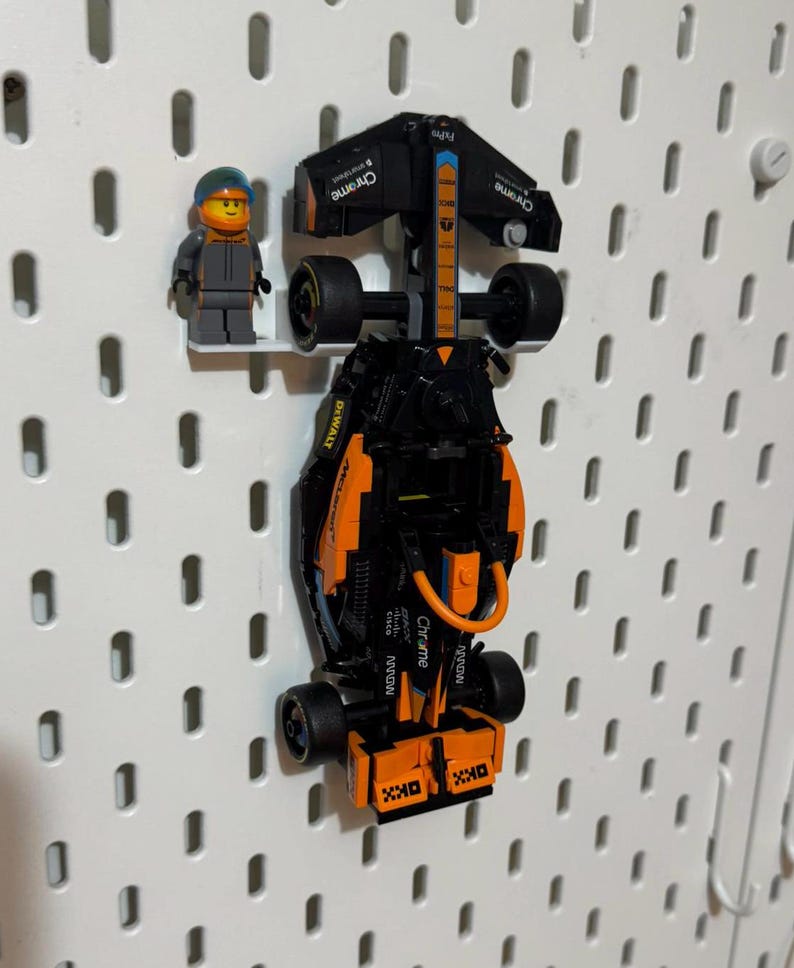 May include: A black and orange Lego Formula 1 race car with the word "Chrome" printed on the side. The car is hanging on a white pegboard. A Lego figure wearing a blue helmet and a grey jumpsuit is standing next to the car.