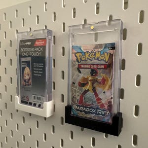 May include: Two clear plastic card holders mounted on a white pegboard. One holder contains a "Booster Pack One-Touch" card, and the other holds a "Pokémon Trading Card Game" pack with "Paradox Rift" text.