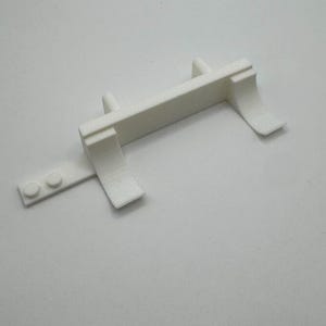 May include: White plastic model of a truck bed with a raised platform and two side rails. The bed has a small, rectangular piece attached to the back.