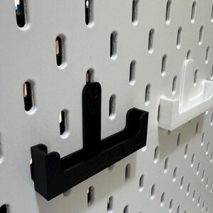 May include: Black and white pegboard with two U-shaped shelf brackets. One bracket is black, the other is white. The pegboard has evenly spaced oval holes. The brackets are designed to hold items on the pegboard.