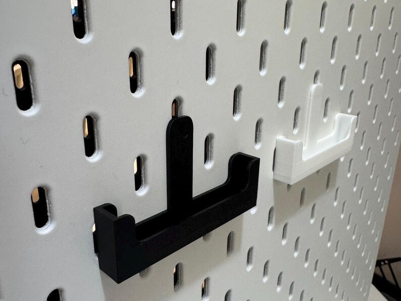 May include: Black and white pegboard with two U-shaped shelf brackets. One bracket is black, the other is white. The pegboard has evenly spaced oval holes. The brackets are designed to hold items on the pegboard.