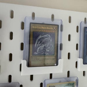 Pu&ograve; includere: Un pannello perforato bianco con un portacarte in plastica trasparente che mostra una carta da gioco "Blue-Eyes White Dragon". La carta &egrave; un'illustrazione blu e bianca di un drago su sfondo dorato. La carta &egrave; circondata da altre carte in portacarte in plastica trasparente.
