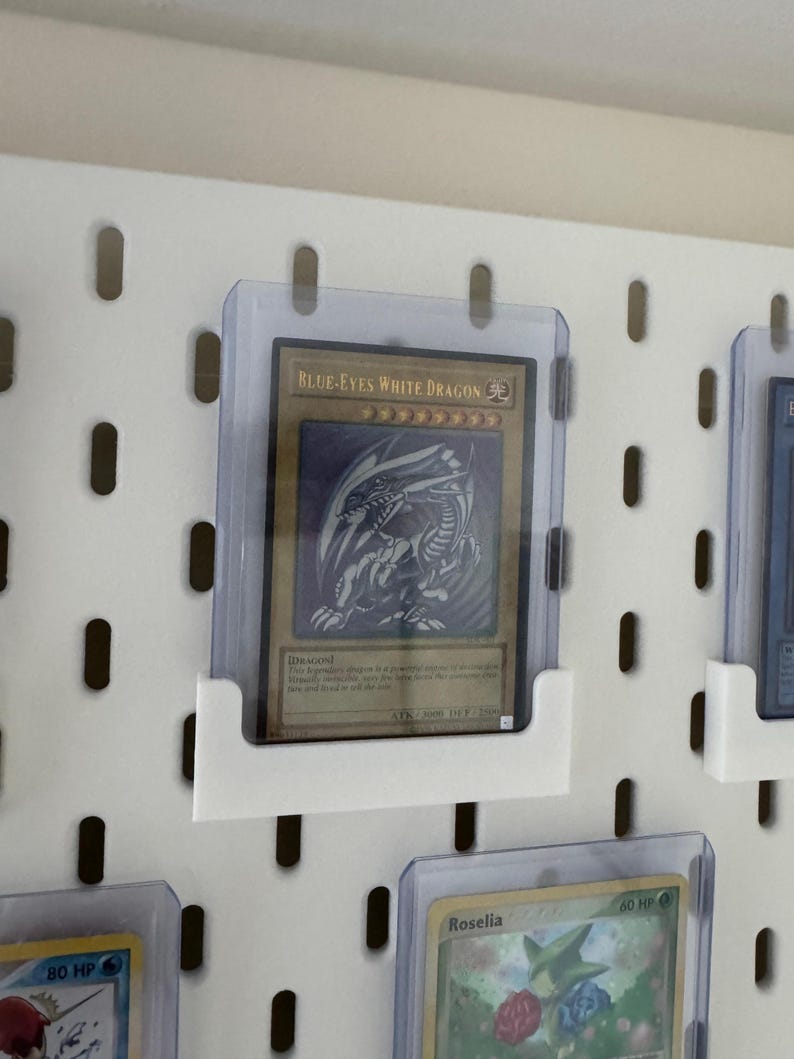 Pu&ograve; includere: Un pannello perforato bianco con un portacarte in plastica trasparente che mostra una carta da gioco "Blue-Eyes White Dragon". La carta &egrave; un'illustrazione blu e bianca di un drago su sfondo dorato. La carta &egrave; circondata da altre carte in portacarte in plastica trasparente.