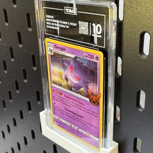 May include: A graded Pok&eacute;mon trading card featuring Gengar from the 2023 Scarlet & Violet set. The card is graded a 10 Gem Mint and is encased in a clear protective holder. The card is displayed on a white stand against a black pegboard.