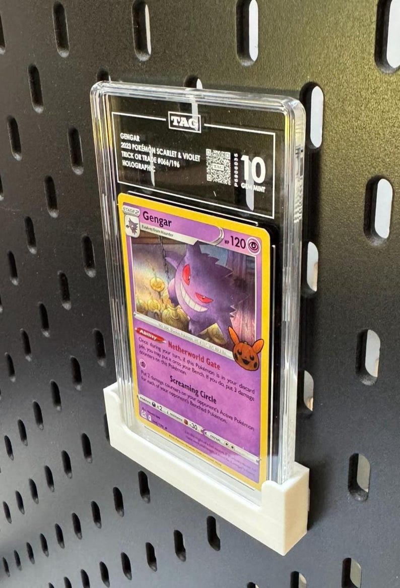 May include: A graded Pok&eacute;mon trading card featuring Gengar from the 2023 Scarlet & Violet set. The card is graded a 10 Gem Mint and is encased in a clear protective holder. The card is displayed on a white stand against a black pegboard.