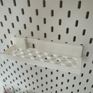 Puede incluir: Una estanter&iacute;a blanca impresa en 3D dise&ntilde;ada para una panel perforado. La estanter&iacute;a tiene forma rectangular con un patr&oacute;n de panal en la base. Est&aacute; montada en un panel perforado blanco con agujeros ovalados espaciados.