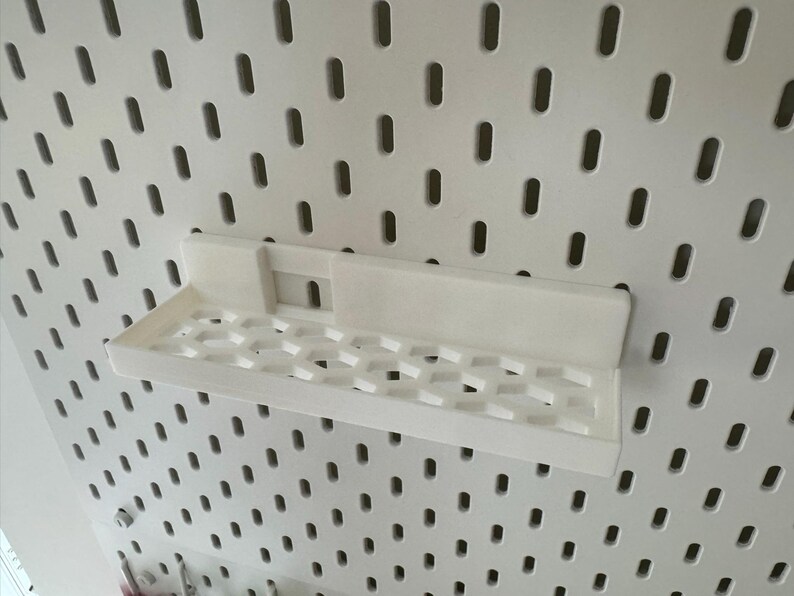 Puede incluir: Una estanter&iacute;a blanca impresa en 3D dise&ntilde;ada para una panel perforado. La estanter&iacute;a tiene forma rectangular con un patr&oacute;n de panal en la base. Est&aacute; montada en un panel perforado blanco con agujeros ovalados espaciados.