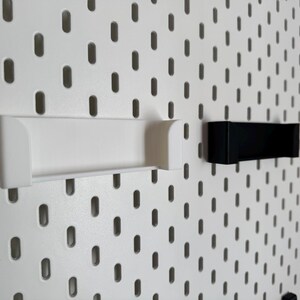 K&ouml;nnte beinhalten: Wei&szlig;e und schwarze Lochwand-Zubeh&ouml;rteile. Das wei&szlig;e Zubeh&ouml;r ist ein langes Regal mit gebogenen Seiten. Das schwarze Zubeh&ouml;r ist ein kleiner, rechteckiger Halter. Beide sind an einer wei&szlig;en Lochwand mit ovalen L&ouml;chern befestigt.
