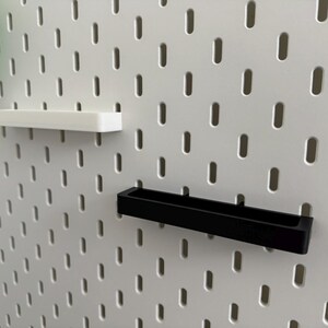 May include: White pegboard with a series of evenly spaced oval holes. Two shelves are attached: a white rectangular shelf and a black rectangular shelf. A green object is partially visible on the left.