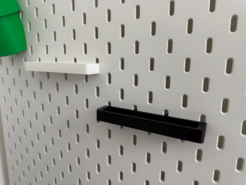 May include: White pegboard with a series of evenly spaced oval holes. Two shelves are attached: a white rectangular shelf and a black rectangular shelf. A green object is partially visible on the left.