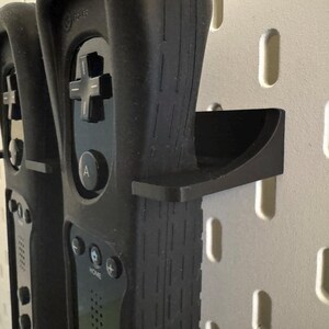 May include: Black Wii remotes with a black holder are mounted on a white pegboard. The remotes have a directional pad, buttons, and a wrist strap. The holder is a curved shelf.