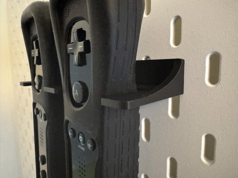 May include: Black Wii remotes with a black holder are mounted on a white pegboard. The remotes have a directional pad, buttons, and a wrist strap. The holder is a curved shelf.