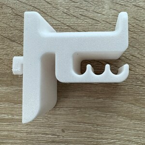 Pu&ograve; includere: Un oggetto in plastica bianca stampato in 3D, forse un supporto o una staffa, con una superficie strutturata. Presenta un design ad angolo retto con un gancio sporgente e tre fessure curve. L'oggetto &egrave; posto su una superficie di legno marrone chiaro.