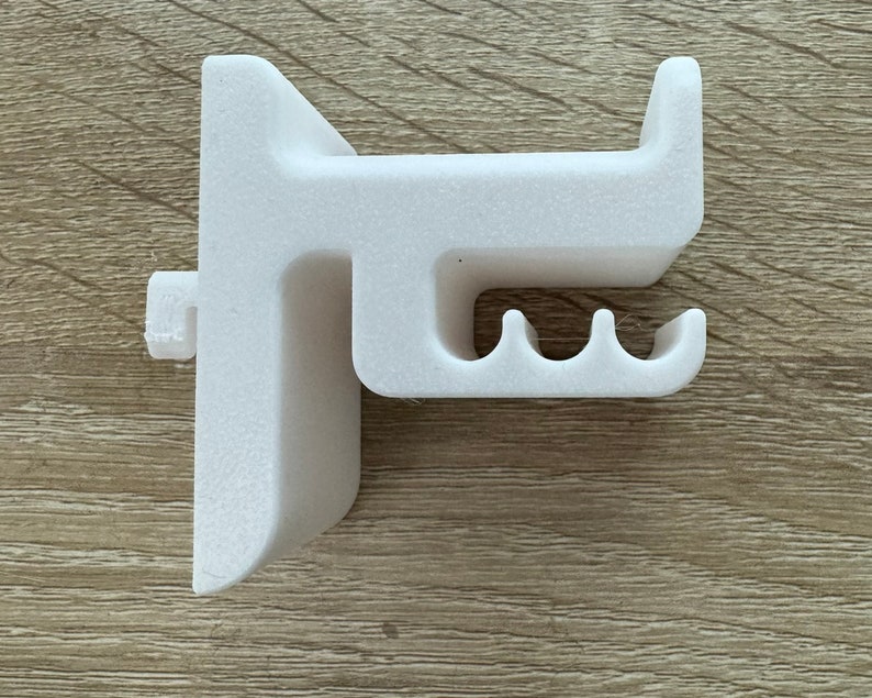 Pu&ograve; includere: Un oggetto in plastica bianca stampato in 3D, forse un supporto o una staffa, con una superficie strutturata. Presenta un design ad angolo retto con un gancio sporgente e tre fessure curve. L'oggetto &egrave; posto su una superficie di legno marrone chiaro.