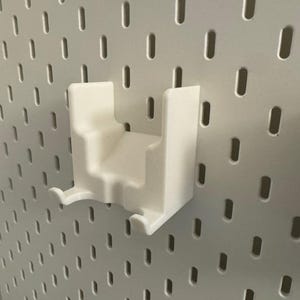 K&ouml;nnte beinhalten: Ein wei&szlig;es 3D-gedrucktes Zubeh&ouml;r f&uuml;r Lochw&auml;nde. Der Artikel hat eine rechteckige Form mit einem abgestuften Design und zwei gebogenen Haken. Es ist an einer wei&szlig;en Lochwand mit gleichm&auml;&szlig;ig verteilten ovalen L&ouml;chern befestigt.