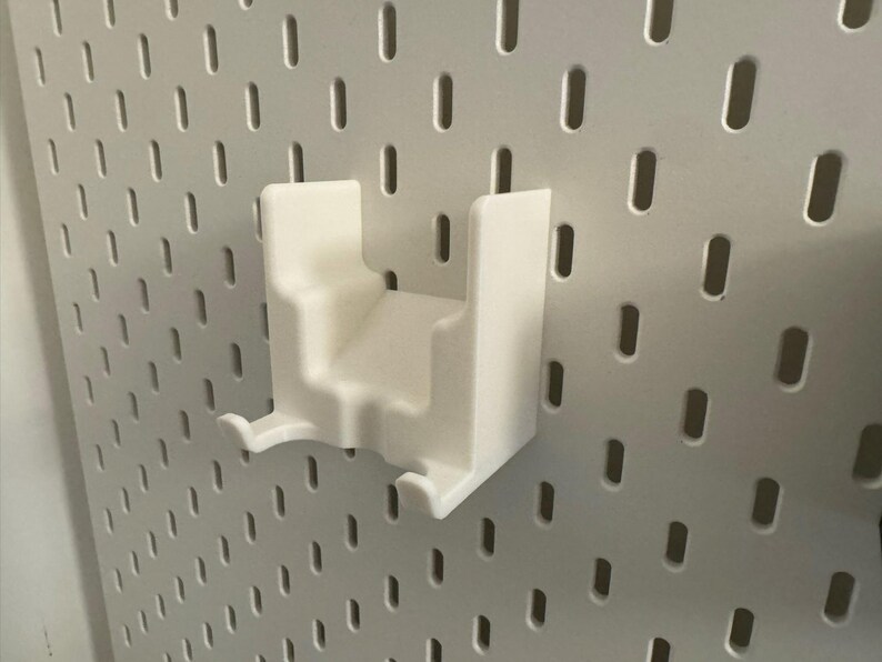 K&ouml;nnte beinhalten: Ein wei&szlig;es 3D-gedrucktes Zubeh&ouml;r f&uuml;r Lochw&auml;nde. Der Artikel hat eine rechteckige Form mit einem abgestuften Design und zwei gebogenen Haken. Es ist an einer wei&szlig;en Lochwand mit gleichm&auml;&szlig;ig verteilten ovalen L&ouml;chern befestigt.