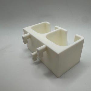 Puede incluir: Un objeto blanco impreso en 3D con dos compartimentos abiertos y dos pesta&ntilde;as rectangulares que sobresalen. El objeto tiene un dise&ntilde;o geom&eacute;trico simple y parece estar hecho de un material s&oacute;lido y opaco. El art&iacute;culo es probablemente un contenedor o soporte.