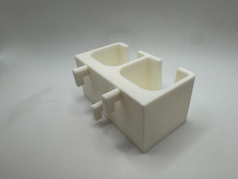 Puede incluir: Un objeto blanco impreso en 3D con dos compartimentos abiertos y dos pesta&ntilde;as rectangulares que sobresalen. El objeto tiene un dise&ntilde;o geom&eacute;trico simple y parece estar hecho de un material s&oacute;lido y opaco. El art&iacute;culo es probablemente un contenedor o soporte.