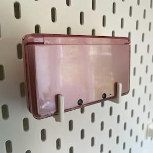 Puede incluir: Una consola de juegos port&aacute;til Nintendo 3DS rosa montada en una pared perforada blanca con soportes blancos. La consola est&aacute; cerrada, mostrando la parte trasera con dos lentes de c&aacute;mara. La pared perforada tiene un patr&oacute;n de agujeros ovalados.