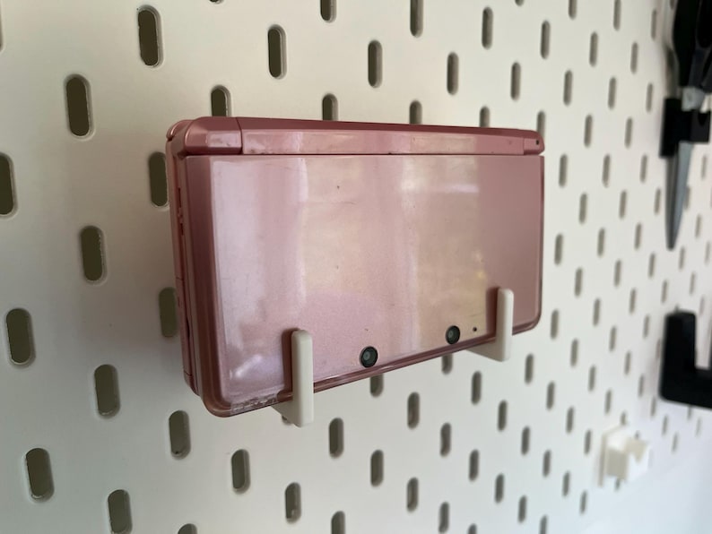 Puede incluir: Una consola de juegos port&aacute;til Nintendo 3DS rosa montada en una pared perforada blanca con soportes blancos. La consola est&aacute; cerrada, mostrando la parte trasera con dos lentes de c&aacute;mara. La pared perforada tiene un patr&oacute;n de agujeros ovalados.