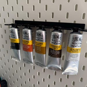 May include: Five tubes of Winsor & Newton Galeria acrylic paint in various colours are displayed on a black metal rack. The tubes are silver with yellow and black labels, mounted on a white pegboard.